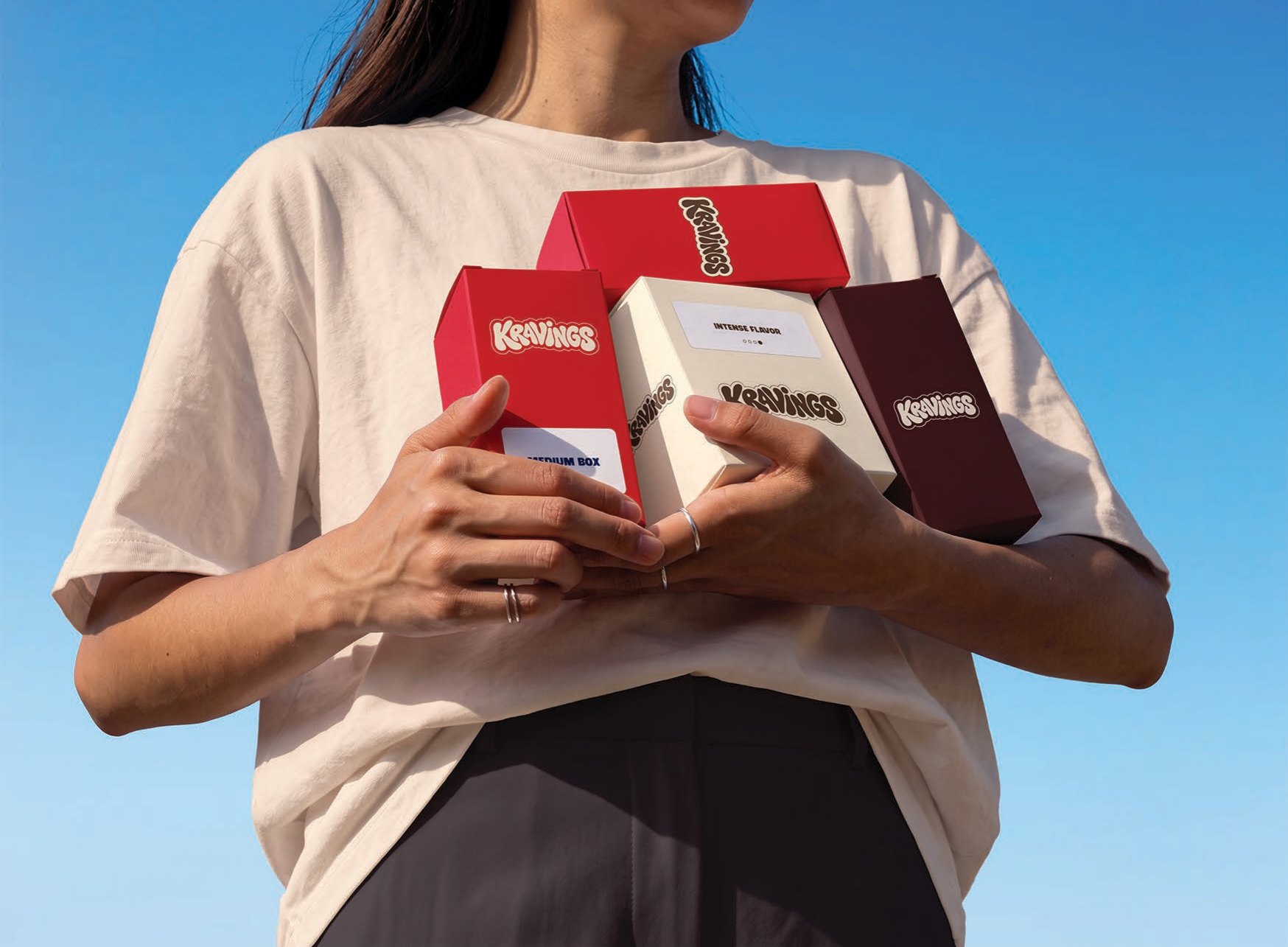 Hands holding red, vanilla and chocolate Kravings boxes against a blue sky